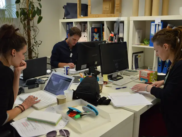 There are 2 women and a man sitting in the chair and doing their work. On the table there are...