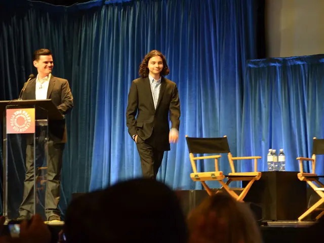 There are two people on a stage one is smiling and standing at the podium, another one is walking....